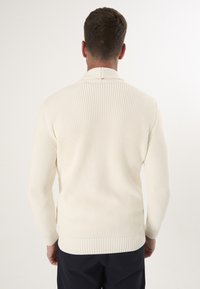 Mees, kes kannab kreemivärvi kudumisse riidejärgset kampsunit ja tumedaid pükse, seisab seljaga tavalise valge tausta poole.