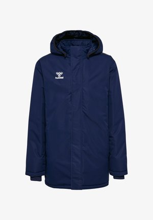 To børn iført marineblå jakker og matchende bukser, stående med armene spredt ud mod en ensfarvet baggrund.