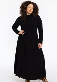 Robe noire longue avec un col montant, des manches longues et un drapé sur le côté ; texture lisse ; portée avec des bottines à imprimé animal.
