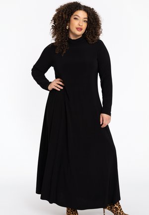 Langes schwarzes Kleid mit hochgeschlossenem Hals, langen Ärmeln und seitlicher Drapierung; glatte Textur; getragen mit Ankle-Boots im Tierprint.