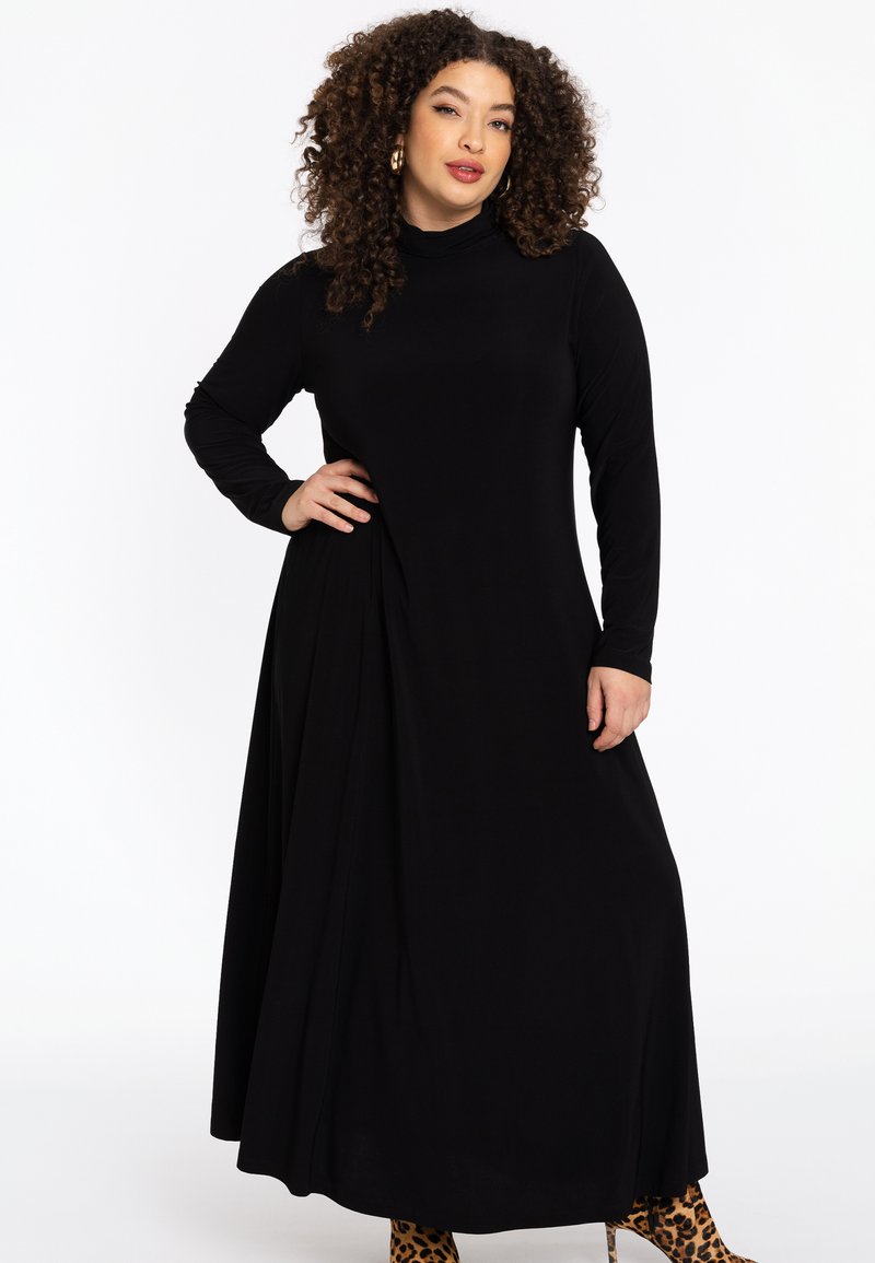 Robe noire longue avec un col montant, des manches longues et un drapé sur le côté ; texture lisse ; portée avec des bottines à imprimé animal.