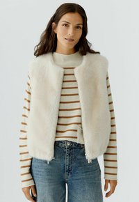 Crème nepbont gilet met een ronde halslijn, gecombineerd met een beige gestreepte trui en denim jeans. Heeft een voorrits en zijzakken.