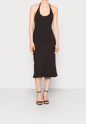 Femme portant une robe midi noire côtelée sans manches à col halter avec des sandales à talons hauts noires à bride à la cheville sur un fond clair uni.