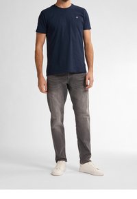 Camiseta de algodón azul marino con cuello redondo y pequeño logo en el pecho, combinada con vaqueros gris y zapatillas blancas.