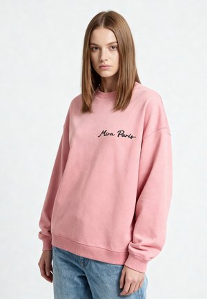 Jeune femme aux cheveux bruns raides portant un sweat rose ample avec le texte « Mira Paris » et un jean bleu, regardant devant avec une expression neutre.