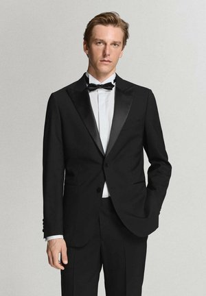 Hombre de pie con esmoquin negro con solapas de satén, camisa blanca y pajarita negra sobre un fondo claro y liso.