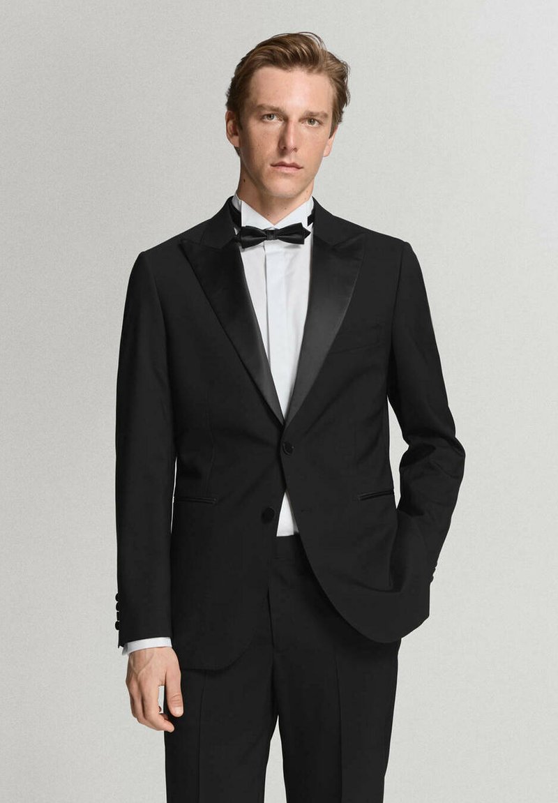 Hombre de pie con esmoquin negro con solapas de satén, camisa blanca y pajarita negra sobre un fondo claro y liso.