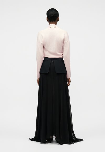Kobieta o ciemnej karnacji i włosach związanych w kok nosi bladoróżową bluzkę z długim rękawem oraz czarną, długą do ziemi spódnicę z detalem panelu z tyłu.