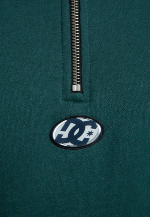 Sudadera de tejido verde azulado con media cremallera que presenta una cremallera metálica y un logotipo ovalado en blanco y negro en el pecho. Textura suave, diseño casual.