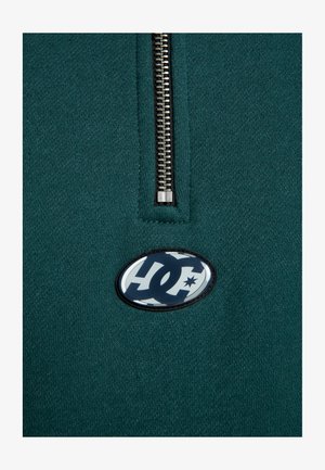 Teal fabric quarter-zip pullover featuring a metal zipper and a black and white oval logo on the chest. Smooth texture, casual design.