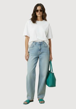 Femme portant un t-shirt blanc oversize et un jean bleu clair, tenant un sac à main bleu sarcelle, portant des sandales bleu sarcelle et des lunettes de soleil, debout devant un fond uni.