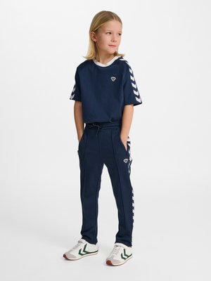 Blondes Kind steht mit den Händen in den Taschen, trägt marineblaue Sportbekleidung mit weißen Chevron-Streifen und weißen Sneakers mit grünen Akzenten.