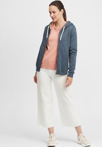 Sweat-shirt à capuche zippé en tissu bleu foncé avec des cordons en crème, associé à un haut rose clair et un pantalon large blanc.