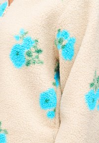 Μπεζ ύφασμα φλις με υφή επιφάνεια διακοσμημένη με διάσπαρτα μπλε και πράσινα λουλουδένια σχέδια.