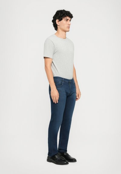 Mlad muškarac u sivoj majici kratkih rukava, tamnoplavim trapericama i crnim cipelama stoji bočno uz jednostavnu bijelu pozadinu.