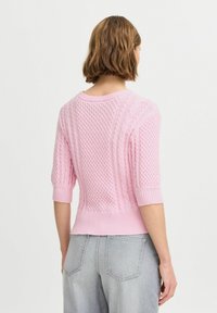 Rosa gestrickter Pullover mit kurzen Ärmeln, der mit strukturierten Mustern und einem gerippten Saum versehen ist. Getragen über hellgrauer Denim.
