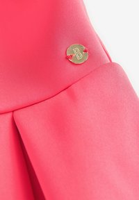 Różowy materiał o gładkiej fakturze, z fałdami i okrągłym złotym akcentem z literą "B" przymocowaną czerwoną nicią.