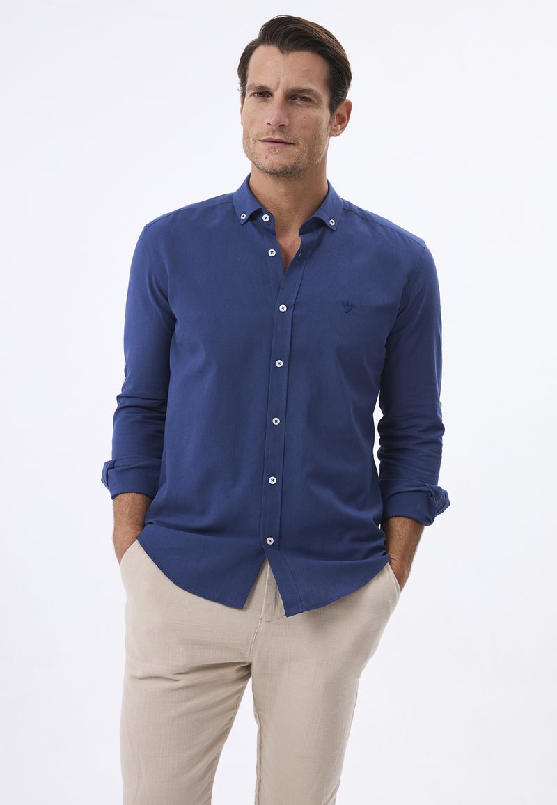 Camisa azul de botones, confeccionada con una tela suave, que presenta un collar puntiagudo y mangas enrolladas. Botones blancos y un logo sutil acentúan el diseño.