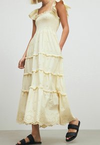Robe maxi en jaune clair avec des volants en cascade, des motifs en œillets et un corsage smocké. Associée à des sandales noires à lanières.