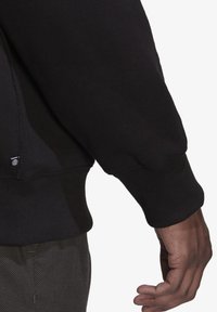 Svart sweatshirt med ribbade muddar, en strukturerad finish och en dragkedjedetalj på sidan, visad på en modells arm och hand.