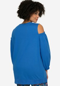 Blå pullovertröja med kallaxel och långa ballongsärmar, tillverkad av mjuk tyg, med avslappnad passform och ribbat nederkant. 