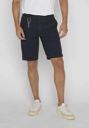 Man die marineblauwe, opgerolde shorts, witte sneakers en een wit T-shirt draagt, staand tegen een effen lichte achtergrond.
