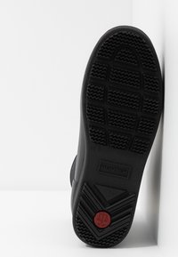 Semelle en caoutchouc noir d'une botte Hunter montrant le motif de la bande de roulement et un emblème circulaire rouge avec un symbole de balances près du talon.