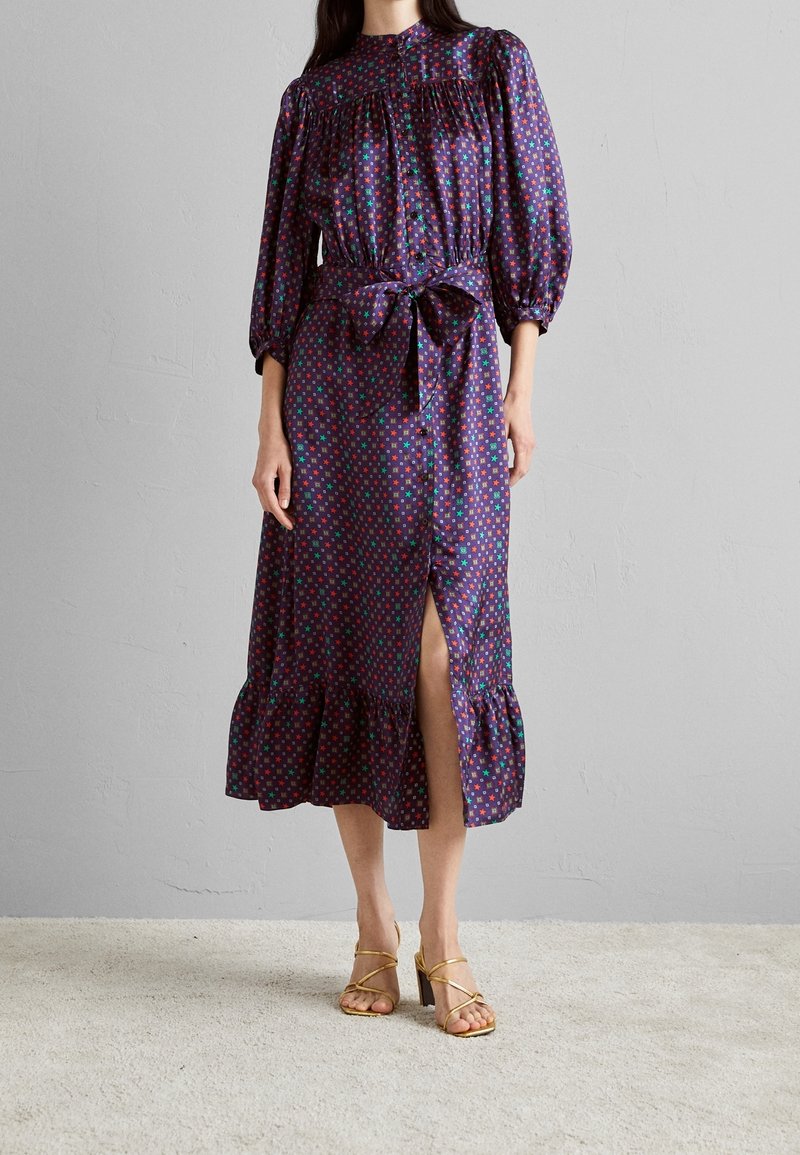 Femme portant une robe midi violette avec des motifs d’étoiles rouges et vertes, manches longues, fente devant, taille nouée et ourlet à volants, associée à des talons dorés.