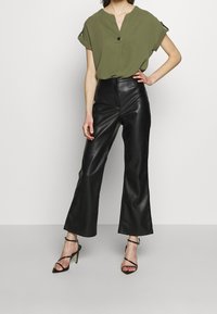 Blusa de manga corta color verde oliva con escote en V y detalle de botones, combinada con pantalones de cuero sintético negros de talle alto y campana, y tacones negros con tiras.