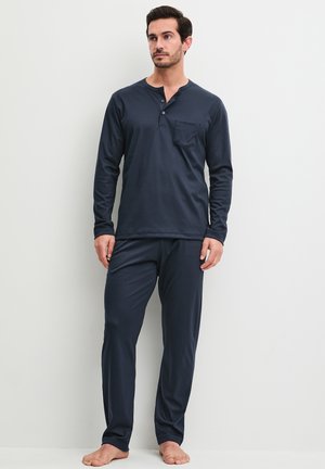 Man staat op blote voeten, draagt een donkerblauw Henley-shirt met lange mouwen en borstzakje en bijpassende broek tegen een effen witte achtergrond.