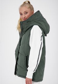 Gilet imbottito verde con colletto alto, tasche con zip e cordini. Indossato sopra una maglietta bianca a maniche lunghe con dettagli neri a fantasia.