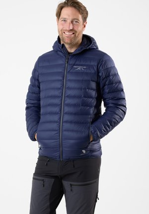 Marineblå puffy jakke med lynlåsåbning foran, tætsiddende hætte og quiltet design. Har logo på brystet og elastiske manchetter. Båret over mørke bukser.