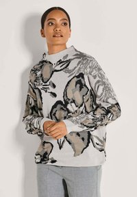 Frau in einem grau-beigen blumigen Musterpullover und hellgrauen Hosen, die mit leicht gefalteten Händen dasteht und zur Seite schaut.