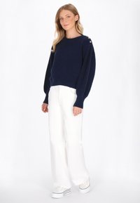 Marineblauwe gebreide trui met een ronde hals en decoratieve knopen op de schouder, gecombineerd met witte wijde broeken en witte sneakers.
