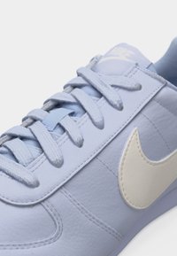 Ανοιχτό μπλε αθλητικό παπούτσι Nike με λευκό λογότυπο swoosh, υφασμένα κορδόνια και ορατές ραφές σε μαλακό δερμάτινο επάνω μέρος.