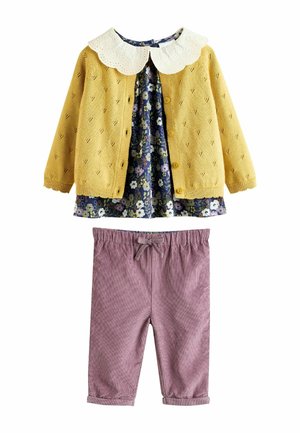 Gelbe Strickjacke mit einem floral bedruckten Kleid darunter, das einen weißen Spitzenkragen hat. Kombiniert mit weichen rosa Cordhosen.