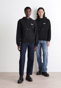 Zwei Modelle stehen nebeneinander und tragen schwarze Kapuzenpullis mit Kordeln und Fronttaschen, kombiniert mit dunkel und hell gewaschenen Jeans sowie schwarzen Stiefeln.