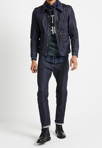 Mörk denimjacka med strukturerad design, parat med mörkblå jeans och svarta snörstövlar. Under jackan en grön sweatshirt med vit text.