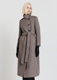 Long manteau gris avec col montant, taille ceinturée et trois boutons. Tissu texturé avec deux poches latérales. Porté avec des gants noirs.