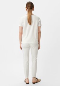 Witte katoenen t-shirt gecombineerd met op maat gemaakte witte broek en beige instappers. Het model heeft lang haar dat in een paardenstaart is gestyled.