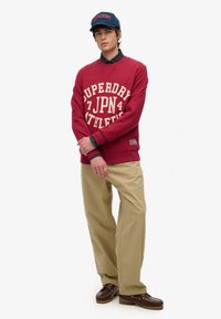 Czerwona bluza z białym napisem "SUPERDRY ATHLETICS", khaki spodnie, ciemne buty i granatowy czapka. Nałożona na koszulę w kratę.