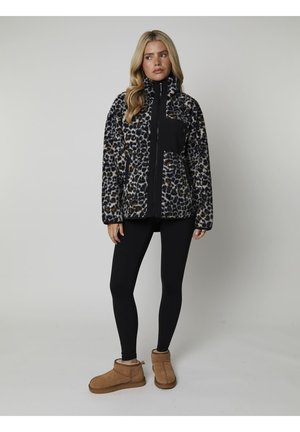 Giacca leopardata nera e beige con colletto alto, chiusura con zip e tasca nera. Indossata sopra leggings neri e stivali color sabbia.