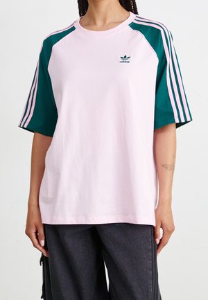 Osoba nosząca jasnoróżową koszulkę Adidas z ciemnozielonymi rękawami, na których znajdują się trzy jasnoróżowe paski oraz logo Adidas na piersi.