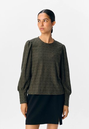 Grønn øyelet genser med lange, puffete ermer, rund halslinning og teksturert mønster. Kombinert med et svart skjørt med sideslitt.