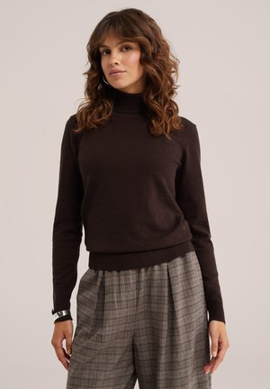 Femme aux cheveux bruns bouclés portant un pull à col roulé noir et un pantalon gris à carreaux taille haute, debout devant un fond uni.