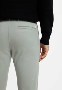 Jasnoszare spodnie o gładkiej teksturze, z tylną kieszenią i małą metką z logo. Ręka modela spoczywa na biodrze.