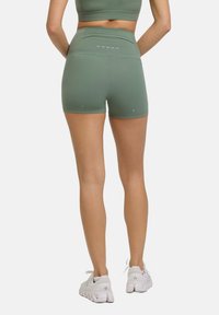Pantaloni sportivi alti in vita di colore verde, con una texture liscia, caratterizzati da cuciture sottili e dettagli bianchi. Abbinati a sneaker bianche.