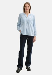 Himmelbluse mit langen Ärmeln und V-Ausschnitt, zwei Brusttaschen und dezenten vertikalen Streifen, kombiniert mit dunkelblauen Schlagjeans und klobigen schwarzen Schuhen.