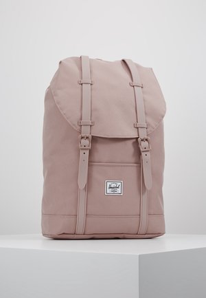 Sac à dos Herschel rose clair avec poche avant et deux sangles à boucle, présenté sur un socle blanc sur fond uni.