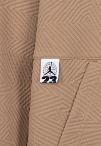 Beige Stoff mit einem strukturierten Fischgrätenmuster und einem weißen Etikett, auf dem die schwarze Silhouette eines Spielers und die Zahl "23" in fetter Schrift zu sehen ist.